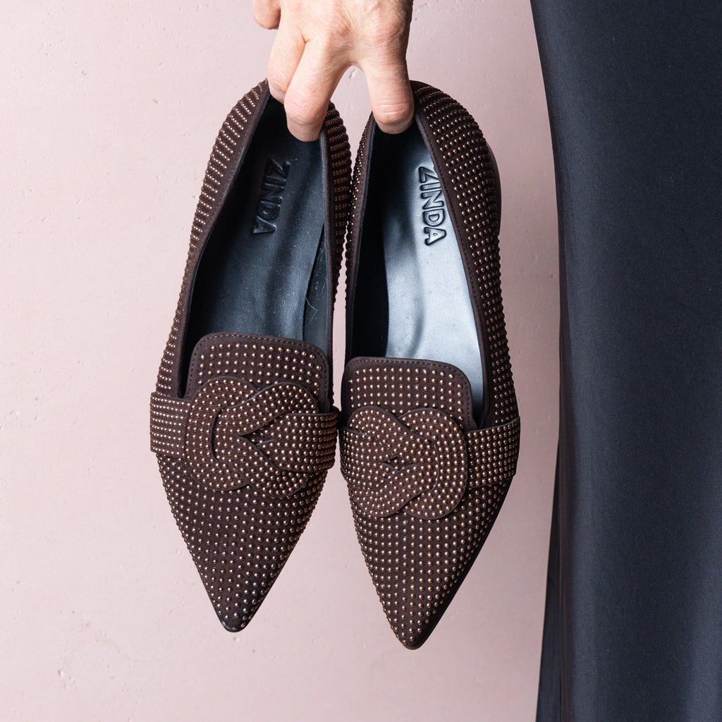 Hand holding a pair of dark brown suede loafers covered in bronze studs in front of a pale pink wall