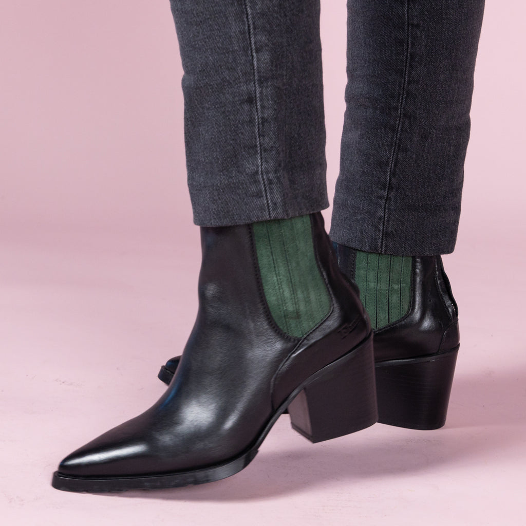 Pair of Black western style boots with a green suede gusset. Woman standing on pink background.