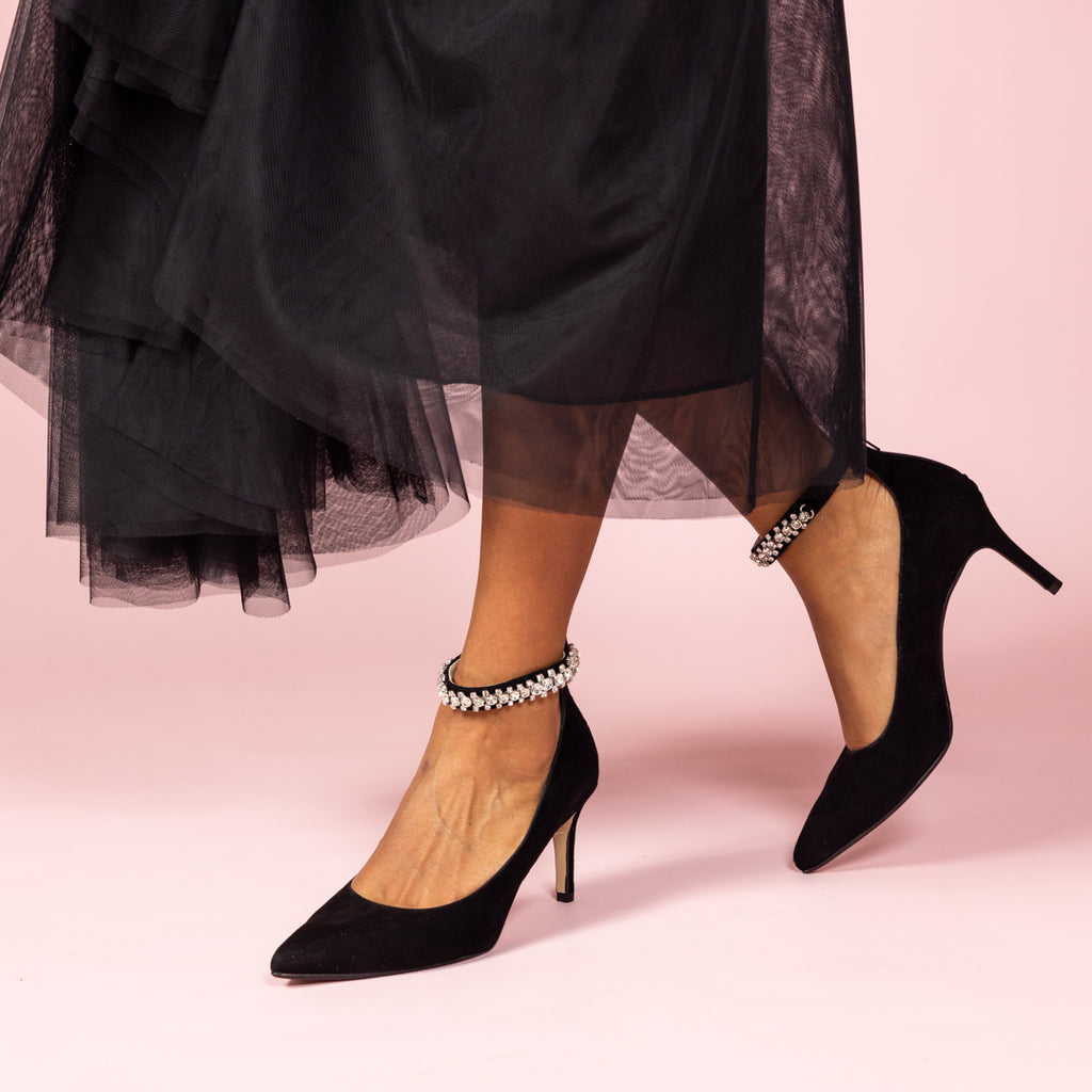 Woman wearing black suede heels with diamante ankle strap and black tulle skirt on pink background