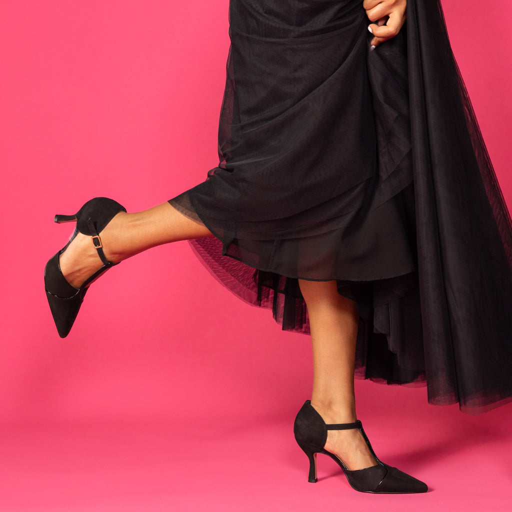 Woman wearing black tutu with t-bar black suede pointed heels kicking up her leg to the back.  On a dark pink background