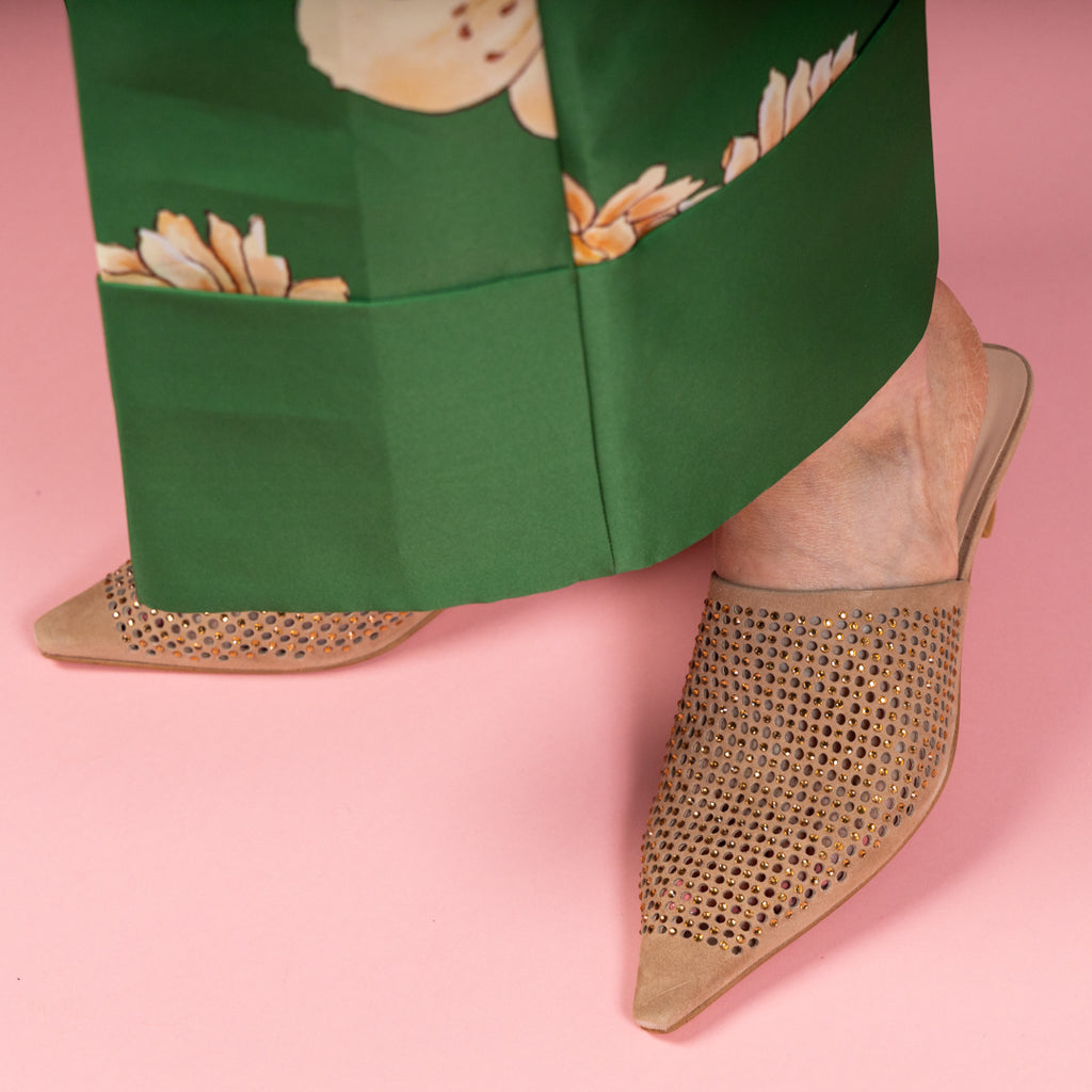 Beige pointed-toe shoes with rhinestone detailing worn with a green floral skirt on a pink background